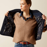 Woman holding a floral-patterned jacket over her shoulders, wearing a brown sweater and blue jeans.